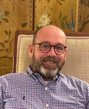 Dr. Jordon Barkalow sitting in a chair smiling with balding light brown hair, a beard and mustache wearing brown rimmed glasses and a blue checked button down shirt